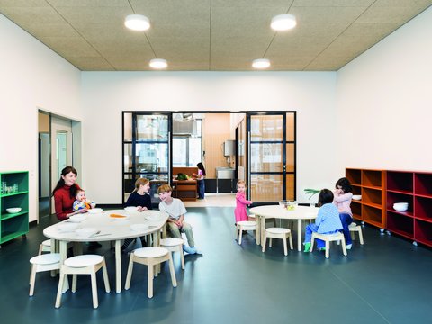 "Großzügiger Kinderbetreuungsraum mit offener Raumstruktur. Links grüne Wandregale mit Utensilien, rechts rot-orange Wandregale. Zwei runde Tische mit beige-weißen Stühlen in der Mitte und rechts. Schwarze Rahmentüren mit Blick in angrenzende Bereiche. Mehrere Kinder und eine Betreuungsperson verteilt im Raum bei verschiedenen Aktivitäten. Warme Beleuchtung, dunkeltürkiser Bodenbelag."