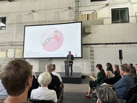 Redner am Podium vor einer Präsentationsleinwand bei einer Veranstaltung in einer Industriehalle.