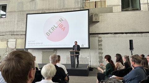 Redner am Podium vor einer Präsentationsleinwand bei einer Veranstaltung in einer Industriehalle.