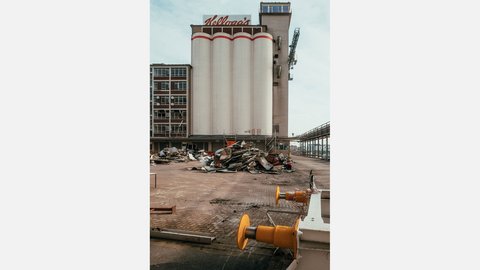 Werksgelände mit hohen weißen Silotürmen mit rotem „Kellogg’s“-Schriftzug; in der Mitte Haufen aus verbogenem Metall und Bauschutt, rechts gelbe Dockpuffer, links ein Bürotrakt und seitlich ein erhöhter Laufsteg.
