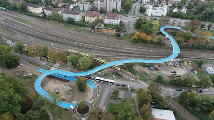 Schlangenförmige Radbrücke aus der Vogelperspektive, eingebettet in Wohngebiet und Verkehrsknoten.