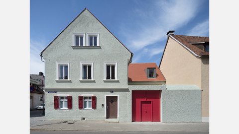 Sanierte historische Hausfassade mit heller Putzoberfläche, roten Fensterläden und farblich abgesetztem Toranbau