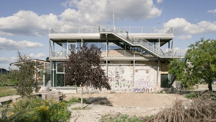 Seitenansicht eines gläsernen Pavillon-Anbaus auf altem Ziegelbau; Außentreppe und Dachterrasse, davor wilder Garten mit Bäumen.