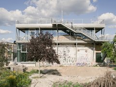 Seitenansicht eines gläsernen Pavillon-Anbaus auf altem Ziegelbau; Außentreppe und Dachterrasse, davor wilder Garten mit Bäumen.