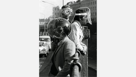 Schwarz-Weiß-Fotografe aus den 1960er Jahren: Zwei Männer tragen futuristische Schutzvorrichtungen mit großen, runden, spiralförmigen Brillengläsern und Kopfbedeckungen. Sie stehen vor einer städtischen Kulisse mit Hochhäusern im Hintergrund. Der Mann links hält eine große metallische Kugel mit spiralförmigen Mustern, der Mann rechts trägt eine flache, rechteckige Vorrichtung. Beide sind in Alltagskleidung gekleidet. Das Bild dokumentiert experimentelle oder künstlerische Visionen von zukünftiger Technologie und Design.