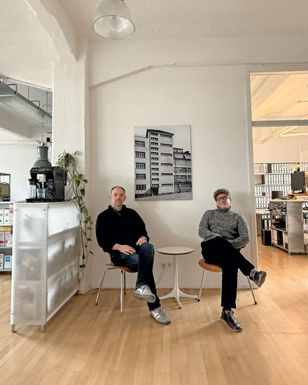Bild 3 (Büro mit zwei Personen): "Heller, minimalistisches Büro mit Holzboden. Zwei Männer sitzen auf weißen Stühlen neben einem kleinen runden Tisch. Links: Mann in schwarzem Pullover und Jeans. Rechts: Mann in grauem Pullover und schwarzer Hose. An der weißen Wand dahinter hängt ein schwarz-weißes Foto eines mehrstöckigen Wohnhauses. Links im Hintergrund eine weiße Möbeleinheit mit einer schwarzen Espressomaschine und hängender grüner Pflanze. Rechts Regale mit Büchern. Von der Decke hängt eine industrielle Leuchte."