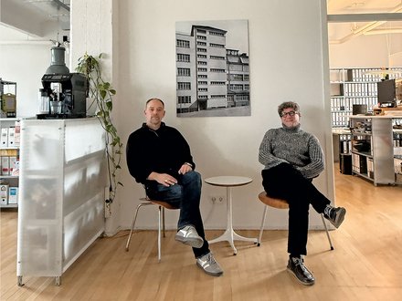 Bild 3 (Büro mit zwei Personen): "Heller, minimalistisches Büro mit Holzboden. Zwei Männer sitzen auf weißen Stühlen neben einem kleinen runden Tisch. Links: Mann in schwarzem Pullover und Jeans. Rechts: Mann in grauem Pullover und schwarzer Hose. An der weißen Wand dahinter hängt ein schwarz-weißes Foto eines mehrstöckigen Wohnhauses. Links im Hintergrund eine weiße Möbeleinheit mit einer schwarzen Espressomaschine und hängender grüner Pflanze. Rechts Regale mit Büchern. Von der Decke hängt eine industrielle Leuchte."