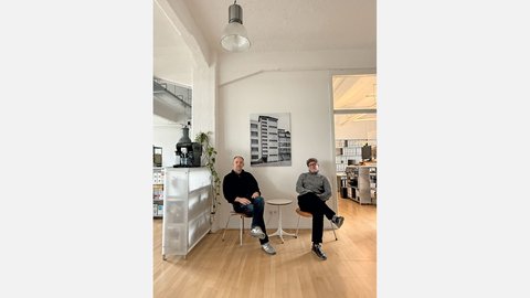 Bild 3 (Büro mit zwei Personen): "Heller, minimalistisches Büro mit Holzboden. Zwei Männer sitzen auf weißen Stühlen neben einem kleinen runden Tisch. Links: Mann in schwarzem Pullover und Jeans. Rechts: Mann in grauem Pullover und schwarzer Hose. An der weißen Wand dahinter hängt ein schwarz-weißes Foto eines mehrstöckigen Wohnhauses. Links im Hintergrund eine weiße Möbeleinheit mit einer schwarzen Espressomaschine und hängender grüner Pflanze. Rechts Regale mit Büchern. Von der Decke hängt eine industrielle Leuchte."