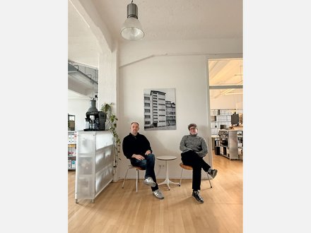 Bild 3 (Büro mit zwei Personen): "Heller, minimalistisches Büro mit Holzboden. Zwei Männer sitzen auf weißen Stühlen neben einem kleinen runden Tisch. Links: Mann in schwarzem Pullover und Jeans. Rechts: Mann in grauem Pullover und schwarzer Hose. An der weißen Wand dahinter hängt ein schwarz-weißes Foto eines mehrstöckigen Wohnhauses. Links im Hintergrund eine weiße Möbeleinheit mit einer schwarzen Espressomaschine und hängender grüner Pflanze. Rechts Regale mit Büchern. Von der Decke hängt eine industrielle Leuchte."