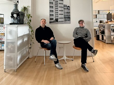 Bild 3 (Büro mit zwei Personen): "Heller, minimalistisches Büro mit Holzboden. Zwei Männer sitzen auf weißen Stühlen neben einem kleinen runden Tisch. Links: Mann in schwarzem Pullover und Jeans. Rechts: Mann in grauem Pullover und schwarzer Hose. An der weißen Wand dahinter hängt ein schwarz-weißes Foto eines mehrstöckigen Wohnhauses. Links im Hintergrund eine weiße Möbeleinheit mit einer schwarzen Espressomaschine und hängender grüner Pflanze. Rechts Regale mit Büchern. Von der Decke hängt eine industrielle Leuchte."