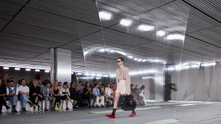 "Ein Model läuft über einen Laufsteg in einem industriellen Innenraum. Durch halbtransparente, herabhängende Folien wirkt die Szene leicht verzerrt. Das Publikum sitzt seitlich und beobachtet die Show. Der Raum ist geprägt von Metalloberflächen und gleichmäßiger Deckenbeleuchtung."