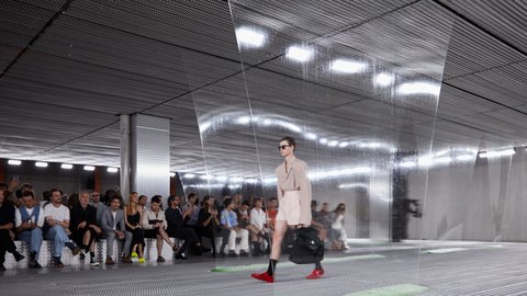 "Ein Model läuft über einen Laufsteg in einem industriellen Innenraum. Durch halbtransparente, herabhängende Folien wirkt die Szene leicht verzerrt. Das Publikum sitzt seitlich und beobachtet die Show. Der Raum ist geprägt von Metalloberflächen und gleichmäßiger Deckenbeleuchtung."