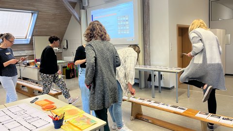 Mehrere Personen stehen und gehen in einem hellen Raum mit Holzbalken, an einer langen Bank liegen Karten oder Papiere, im Hintergrund ist eine Projektionsfläche an der Wand zu sehen.