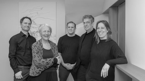 Schwarz-Weiß-Porträt eines fünfköpfigen Teams in einem modernen Büro vor einer künstlerischen Wandzeichnung.