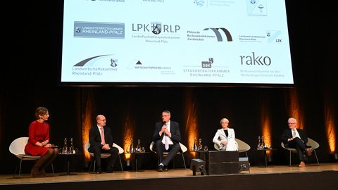 Podiumsdiskussion mit fünf Personen auf der Bühne; die mittlere Person spricht ins Mikrofon. Dahinter große Projektionsfläche mit Logos von Kammern und Verbänden.