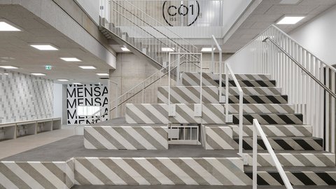 Schulinnenraum: breite Sitztreppe aus Beton mit diagonalen Streifen neben Geländern; Beschriftung „MENSA“ im Hintergrund.