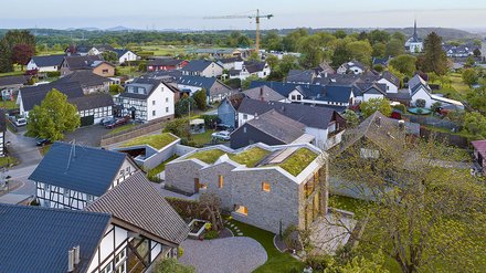 Luftaufnahme eines Dorfes mit gemischter Bebauung. Im Vordergrund (unten links) ein modernes Gebäude mit dunklem Blechdach und Fachwerk-Elementen. Zentral (Bildmitte) ein neues, großes Steingebäude mit grauem Naturstein, Flachdach und extensiver Dachbegrünung mit grüner Rasenfläche. Das Gebäude hat große, warmgelb beleuchtete Fenster und ist von grünen Hecken und Vegetation umgeben. Dahinter erstreckt sich das traditionelle Dorf mit verschiedenen älteren Häusern: Fachwerkhäuser, weiße und braune Fassaden, dunkle Satteldächer. Eine Baustelle mit gelben Baugerüsten ist sichtbar. Im Hintergrund hügeliges grünes Landschaftspanorama mit Feldern, Wiesen und Baumgruppen. Der Himmel ist teilweise bewölkt, die Sonne scheint."