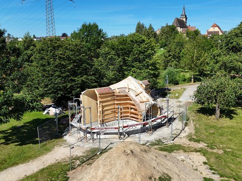 "Baustelle einer Holzkirche im Grünen. Im Vordergrund eine hölzerne Gewölbestruktur unter Baugerüst und Schutzplanen. Umgeben von dichtem Wald, grünen Wiesen und metallischem Stromleitungsmast. Im Hintergrund eine traditionelle Dorfkirche mit spitzem Turm und rote Dachziegel. Klarer blauer Himmel."