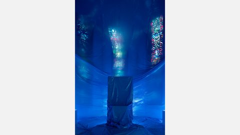Blau beleuchteter Kirchenraum mit verhülltem Objekt und farbig schimmernden Kirchenfenstern als Teil einer Kunstinstallation.