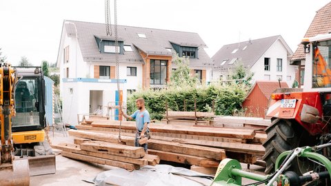 Baustellenhof mit Bagger und Traktor; Person richtet Ketten über gestapelten Altholzbalken aus.