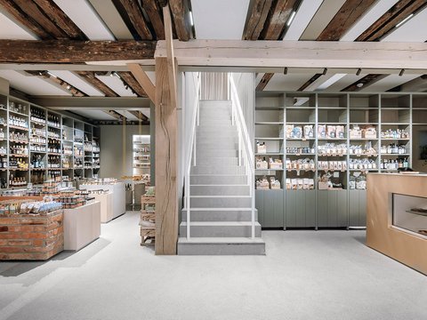 "Moderner Einzelhandelsladen mit hohen Holzbalken an der Decke. Umfassend bestückte Regale mit weißen Produkten an beiden Wänden. Rustikale Backsteinelemente am Tresen links. Zentrale Treppe führt nach oben. Heller, offener Raum mit gemischtem rustikalem und modernem Design."