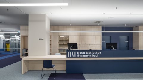 "Moderner Bibliotheks-Ausleihbereich. Großer geschwungener Tresen aus Holz und Dunkelblau mit weißem Logo "Neue Bibliothek Gummersbach" und integrierten Bücherregalen. Zwei Computer-Arbeitsplätze hinter dem Tresen. Links ein Serviceschalter mit Symbol. Heller Bodenbelag mit dunkelblauen Zonen. Mehrere weiße Deckenleuchten. Im Hintergrund Bücherregale und Zugangsbereiche."