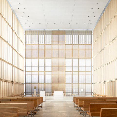 Minimalistischer Innenraum mit Holzbänken links und rechts, große Fensterfront mit Rasterstruktur in Beige- und Weisstönen, heller Betonfußboden, schlicht gestalteter Altar im Zentrum