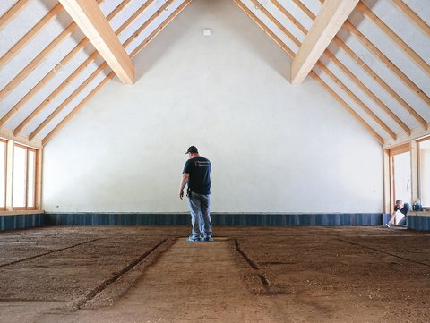 "Ein Mann in schwarzem T-Shirt mit Aufdruck und blauer Jeans steht allein in einer großen, leeren Reithalle mit Holzkonstruktion. Der Raum hat ein beeindruckendes Satteldach mit sichtbaren hellen Holzbalken und weißer Innenwandverkleidung. Der Boden besteht aus dunkelbraunem Reitplatz-Material mit Spuren von Reiten. An den Seiten des Raumes befinden sich großflächige Fenster mit hellen Rahmen, durch die Tageslicht einfällt. Rechts im Hintergrund sitzt eine Person teilweise verborgen in der Ecke. Der Raum wirkt sehr geräumig und hell, mit minimalem Inhalt, was die Größe und Leere der Halle unterstreicht."