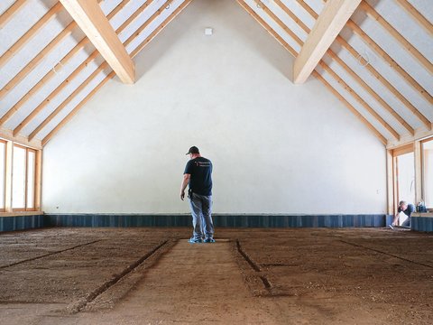 "Ein Mann in schwarzem T-Shirt mit Aufdruck und blauer Jeans steht allein in einer großen, leeren Reithalle mit Holzkonstruktion. Der Raum hat ein beeindruckendes Satteldach mit sichtbaren hellen Holzbalken und weißer Innenwandverkleidung. Der Boden besteht aus dunkelbraunem Reitplatz-Material mit Spuren von Reiten. An den Seiten des Raumes befinden sich großflächige Fenster mit hellen Rahmen, durch die Tageslicht einfällt. Rechts im Hintergrund sitzt eine Person teilweise verborgen in der Ecke. Der Raum wirkt sehr geräumig und hell, mit minimalem Inhalt, was die Größe und Leere der Halle unterstreicht."