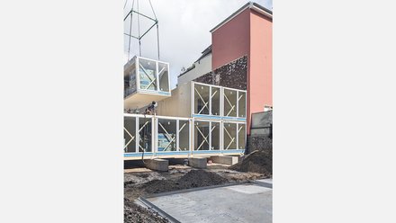 Baustelle: Raummodul wird per Kran auf gestapelte Module gesetzt; Arbeiter auf den Elementen, rosa Brandwand im Hintergrund.