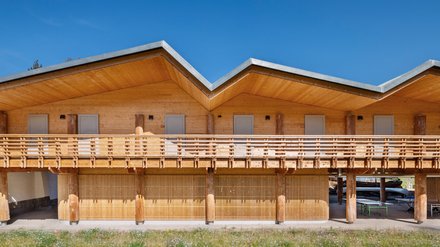 rontansicht des langgestreckten Holzbaus mit gefaltetem Satteldach, Balkonlamellen und Rundholzstützen.