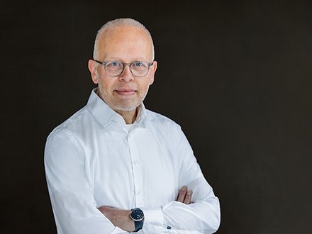 Porträt einer Person mit Brille im weißen Hemd, Arme verschränkt und Armbanduhr; neutraler dunkler Hintergrund, freundlicher Ausdruck.