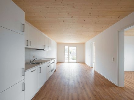 "Offene Wohnküche mit modernem Design. Links an der Wand eine weiße Küchenzeile mit Arbeitsplatte und Kochfeld. Naturholzdecke in heller Farbe. Großer Wohn- und Essbereich mit Holzfußboden. Zwei Glastüren führen auf einen Balkon mit Ausblick. Rechts mehrere weiße Türen zu anderen Räumen. Der Raum ist hell und großzügig geschnitten."
