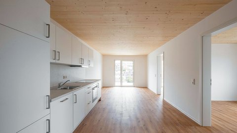 "Offene Wohnküche mit modernem Design. Links an der Wand eine weiße Küchenzeile mit Arbeitsplatte und Kochfeld. Naturholzdecke in heller Farbe. Großer Wohn- und Essbereich mit Holzfußboden. Zwei Glastüren führen auf einen Balkon mit Ausblick. Rechts mehrere weiße Türen zu anderen Räumen. Der Raum ist hell und großzügig geschnitten."