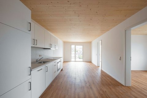 "Offene Wohnküche mit modernem Design. Links an der Wand eine weiße Küchenzeile mit Arbeitsplatte und Kochfeld. Naturholzdecke in heller Farbe. Großer Wohn- und Essbereich mit Holzfußboden. Zwei Glastüren führen auf einen Balkon mit Ausblick. Rechts mehrere weiße Türen zu anderen Räumen. Der Raum ist hell und großzügig geschnitten."