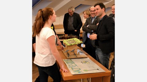 "Ein Mädchen mit einem Pferdeschwanz steht im Vordergrund und schaut zu zwei Männern, die sich angeregt über ein Modell einer Bushaltestelle auf dem Tisch unterhalten. Das Modell hat ein grünes Dach und blaue Bänke. Auf dem Tisch liegt auch ein großes Plakat mit der Aufschrift 'HALTESTELLE', wobei das 'H' als Bushaltestellensymbol gestaltet ist."