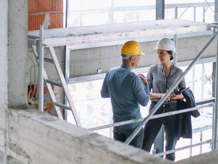 Zwei Personen mit Schutzhelmen unterhalten sich auf einer Baustelle, eine Person hält eine Mappe