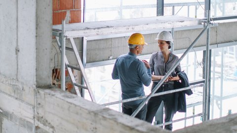 Zwei Personen mit Schutzhelmen unterhalten sich auf einer Baustelle, eine Person hält eine Mappe