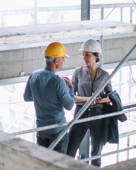 Zwei Personen mit Schutzhelmen unterhalten sich auf einer Baustelle, eine Person hält eine Mappe