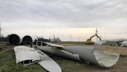 Blick auf eine Baustelle im flachen Land: im Vordergrund liegen mehrere lange, aufgeschnittene und gebrochene Rotorblätter von Windenergieanlagen auf dem Boden, ihre inneren Querschnitte sind sichtbar. Weiter hinten arbeitet ein gelber Bagger an einem der Rotorsegmente, zwei Personen stehen in der Nähe. Am Horizont mehrere noch aufgestellte Windräder, darüber ein bewölkter Himmel.