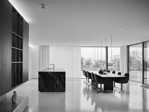 Modernes, minimalistisches Büroinneres in Schwarzweiß. Der Raum zeigt eine offene Küche mit schwarzer Marmorarbeitsplatte und Wasserhahn auf der linken Seite, daneben ein dunkles Regalwandsystem. Im Hintergrund ein großer Konferenztisch mit sechs schwarzen Bürostühlen. Rechts des Raumes befinden sich raumhohe Fenster mit weißen Vorhängen, die den Blick auf kahle Bäume und eine Stadtlandschaft freigeben. Die Decke ist mit indirekter Beleuchtung und hängenden Metallelementen gestaltet. Der Boden ist ein glänzender grauer Estrich. Die Gesamtgestaltung wirkt sehr hell und luftig mit strengem, geometrischem Design.