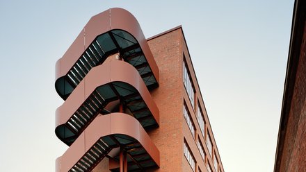 Backsteingebäude mit markanter außenliegender Treppenanlage aus rot lackiertem Stahl.