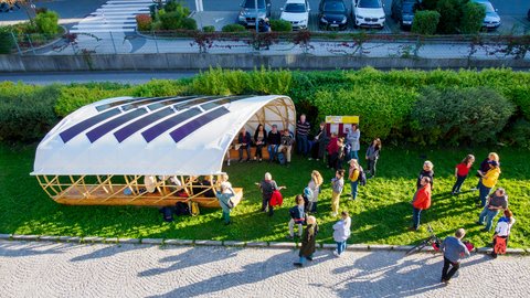 "Blick aus der Vogelperspektive auf eine Menschengruppe, die auf einer Wiese um einen offenen Holzpavillon steht. Auf dem weißen, gebogenen Dach des Pavillons sind dunkle Solarmodule angebracht."