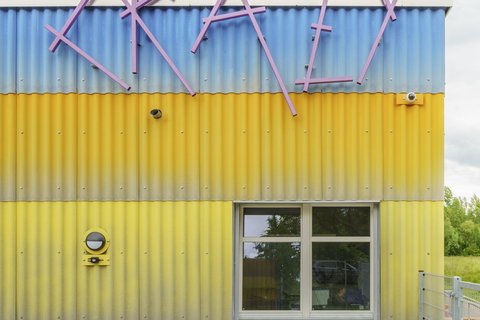 Alt-Text: Detailansicht der farbenfrohen Wellblechfassade des Neubaus. Die Streifen sind lila, gelb und blau. Oben ist der Schriftzug "CRAZY" in großen, unregelmäßigen, lilafarbenen Buchstaben angebracht. Darunter befindet sich ein Fenster. Ein grüner, runder Lautsprecher ist links an der gelben Wand befestigt.