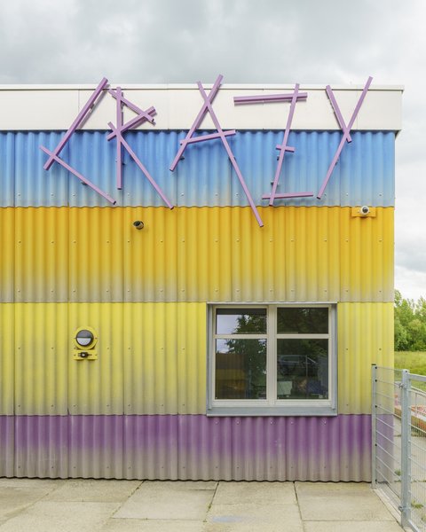 Alt-Text: Detailansicht der farbenfrohen Wellblechfassade des Neubaus. Die Streifen sind lila, gelb und blau. Oben ist der Schriftzug "CRAZY" in großen, unregelmäßigen, lilafarbenen Buchstaben angebracht. Darunter befindet sich ein Fenster. Ein grüner, runder Lautsprecher ist links an der gelben Wand befestigt.