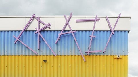 Alt-Text: Detailansicht der farbenfrohen Wellblechfassade des Neubaus. Die Streifen sind lila, gelb und blau. Oben ist der Schriftzug "CRAZY" in großen, unregelmäßigen, lilafarbenen Buchstaben angebracht. Darunter befindet sich ein Fenster. Ein grüner, runder Lautsprecher ist links an der gelben Wand befestigt.