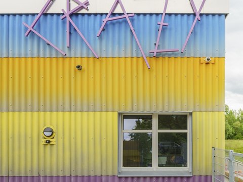 Alt-Text: Detailansicht der farbenfrohen Wellblechfassade des Neubaus. Die Streifen sind lila, gelb und blau. Oben ist der Schriftzug "CRAZY" in großen, unregelmäßigen, lilafarbenen Buchstaben angebracht. Darunter befindet sich ein Fenster. Ein grüner, runder Lautsprecher ist links an der gelben Wand befestigt.