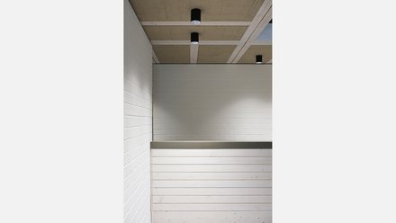 Nahaufnahme der Empfangstheke aus hellem Holz vor weißer Ziegelwand; darüber Rasterdecke mit schwarzen Zylinderleuchten.