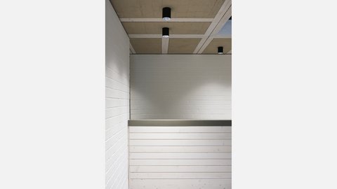Nahaufnahme der Empfangstheke aus hellem Holz vor weißer Ziegelwand; darüber Rasterdecke mit schwarzen Zylinderleuchten.