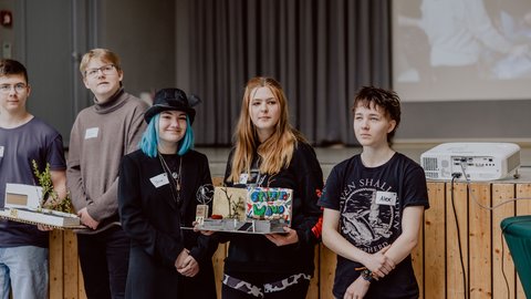 Gruppe von Schülerinnen und Schülern präsentiert ihr Architekturprojekt vor Publikum während einer Abschlussveranstaltung.